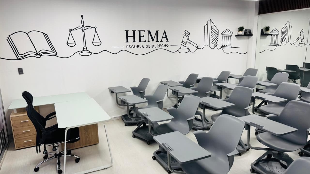 Frente de la misma aula: el escritorio del docente con su silla y un archivero de madera, bajo el mural del libro abierto y la balanza de la justicia; a la derecha, las butacas de los estudiantes.