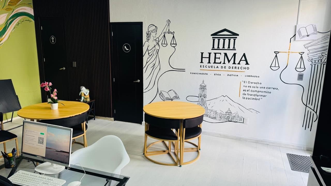 Zona de estudio bajo el mural completo de la escuela: la Justicia con los ojos vendados sosteniendo la balanza, el logotipo «HEMA · Escuela de Derecho» con su frontón de columnas, el Reloj Monumental de Pachuca y la frase «El Derecho no es solo una carrera, es el compromiso de transformar la sociedad»; delante, dos mesas redondas de madera con sillas.
