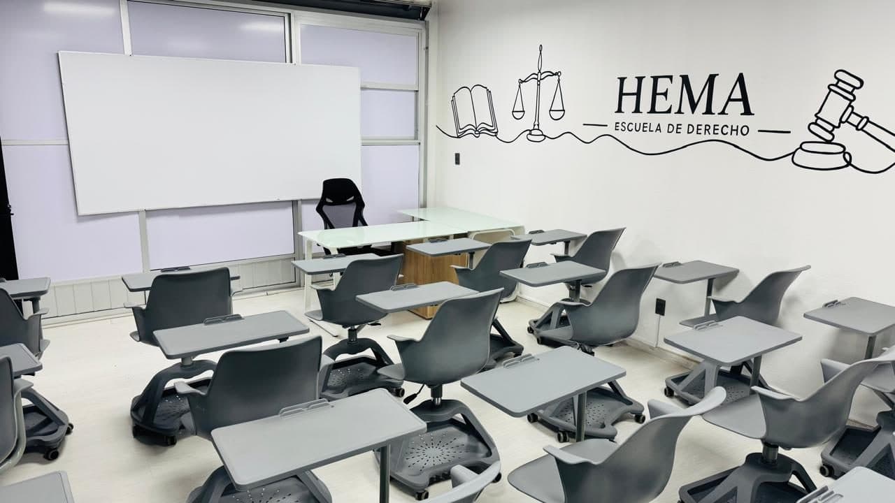 El aula desde la esquina contraria: el pizarrón blanco junto a los ventanales, el escritorio del frente y las butacas grises alineadas en filas.